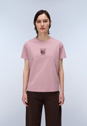 Femme aux cheveux courts et foncés portant un t-shirt rose clair Napapijri Research et un pantalon marron foncé, debout devant un fond clair uni.