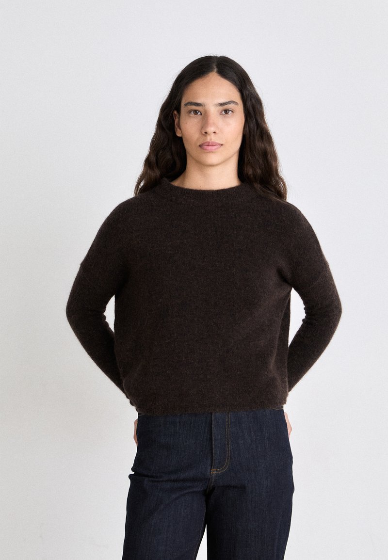Brown, knitted sweater with a round neckline, long sleeves, and soft texture. Paired with dark denim jeans, standing against a neutral background.