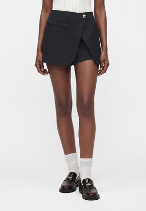 Femme portant un short noir style portefeuille avec un bouton devant, des chaussettes blanches et des mocassins en cuir marron foncé sur fond blanc.
