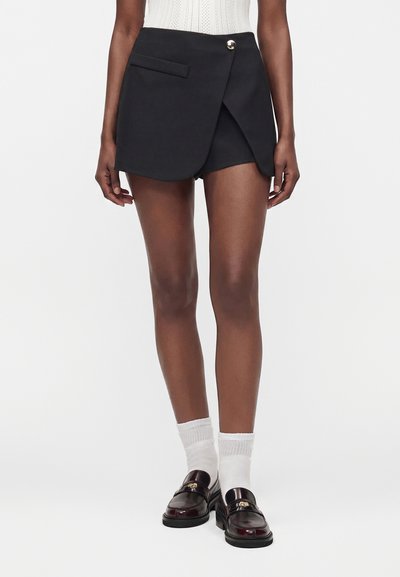 Femme portant un short noir style portefeuille avec un bouton devant, des chaussettes blanches et des mocassins en cuir marron foncé sur fond blanc.