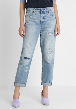 Jasnoniebieskie postrzępione, podwinięte jeansy noszone z liliowymi butami na wysokim obcasie, pokazane od talii do stóp na jednolitym tle.