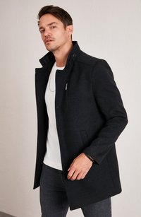 Abrigo de mezcla de lana negra con un cuello estructurado, detalles de botones y bolsillos laterales, combinado con una camisa blanca y jeans oscuros.