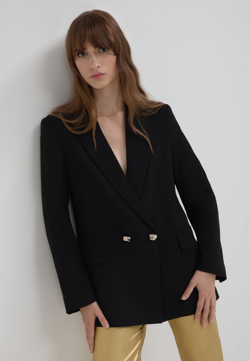 Blazer nero doppiopetto con profondo scollo a V, dettagli in metallo dorato e maniche allungate, abbinato a pantaloni dorati lucidi.