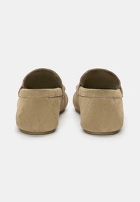 Un paio di mocassini in suede beige visti da dietro, con tacco arrotondato, cuciture visibili e suole piatte in gomma su uno sfondo semplice.