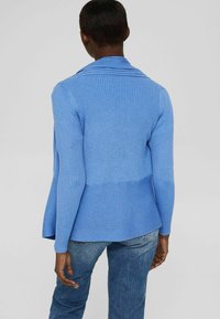 Person som bär en ribbstrukturad blå cardigan med bred krage och blå jeans, sedd bakifrån mot en neutral bakgrund.