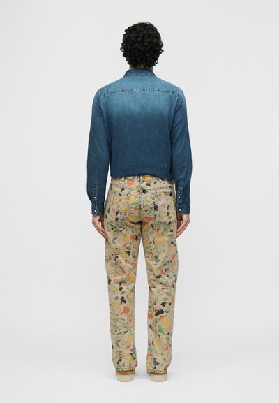 Homme aux cheveux bouclés vu de dos, portant une veste en denim bleu et un pantalon clair avec un motif floral abstrait coloré.