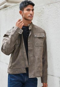 Chaqueta de imitación cuero marrón con cremallera frontal, botones a presión y bolsillos en el pecho. La tela tiene una textura suave y un diseño casual.