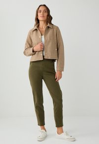 Giacca cropped marrone con colletto a punta, chiusura a bottoni e due tasche frontali, abbinata a pantaloni verde oliva e sneakers bianche.