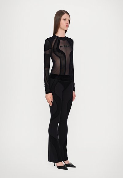 Sheer black long-sleeve top with vertical stripes and logo; high-waisted black pants with textured accents and flared hems. Pointed black heels.