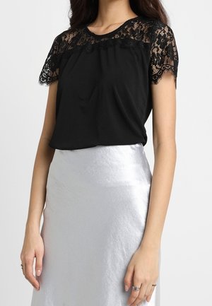 Vrouw draagt een zwart kanten top met korte mouwen, gecombineerd met een zilveren satijnen rok met hoge taille, staand voor een effen achtergrond.