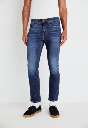 Jean en denim bleu foncé avec une coupe slim, un design à cinq poches, un délavage subtil et des coutures contrastantes. Assorti avec des chaussures foncées et des chaussettes blanches.