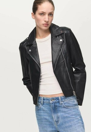 Zwarte leren motorjas met ritssluiting, drukknopenkraag en zilverkleurige details, gedragen over een wit topje en lichtblauwe jeans.