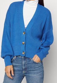 Blå stickad cardigan med djup V-ringning, tre stora knappar, ribbad textur och överdimensionerade ärmar, stylad över en vit polo.