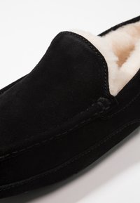 Svart mockaskor med en mjuk beige foder. Har en moccasin-design, rund tå och synlig sömnad längs kanterna.
