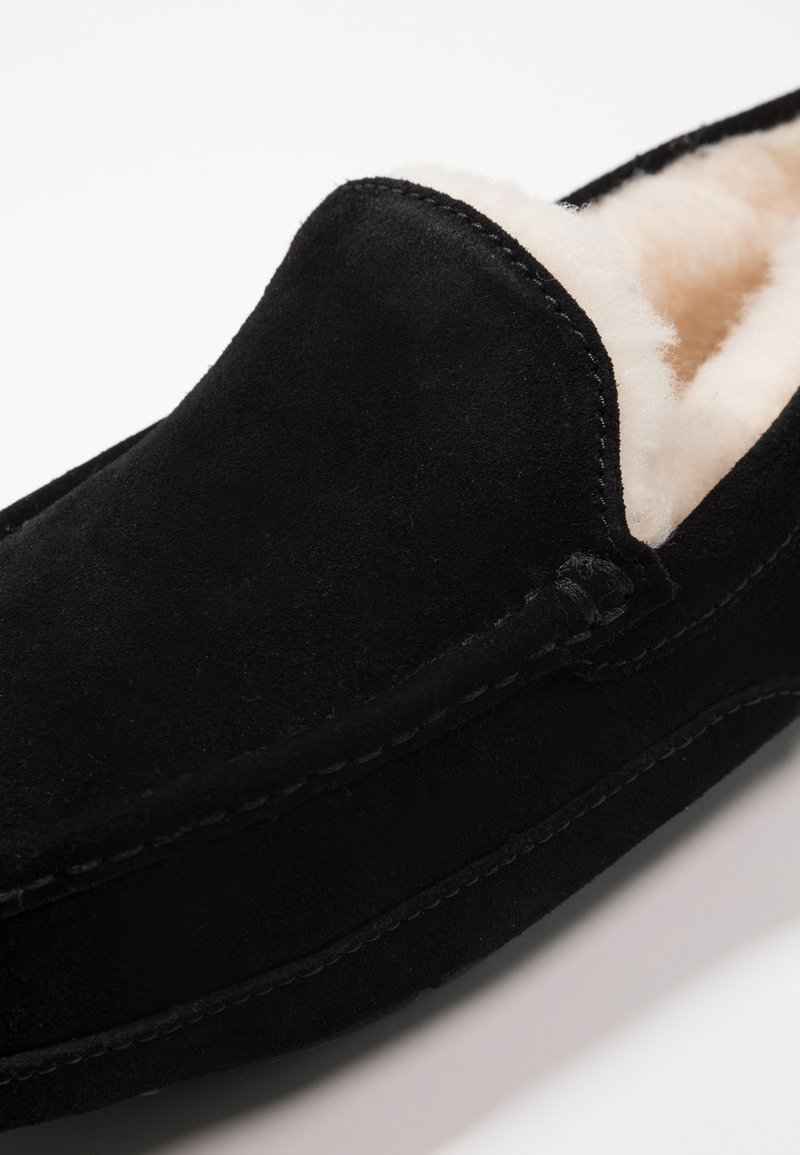 Black suede slipper with a soft beige lining. Features a moccasin design, rounded toe, and visible stitching along the edges.