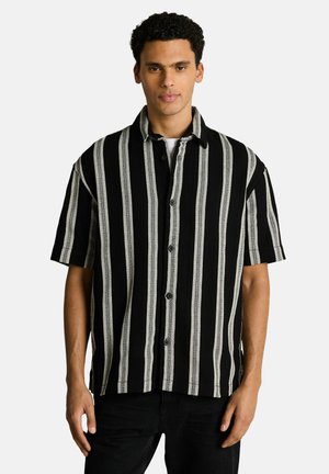 Man wearing short-sleeve black shirt with wide vertical white stripes, standing against plain light background.