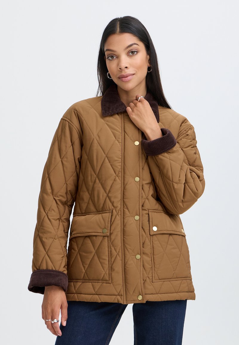 Femme portant une veste matelassée marron avec un col et des poignets en velours côtelé brun foncé, debout devant un fond clair uni.