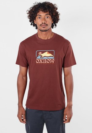 Homme aux cheveux bouclés porte un t-shirt marron avec un motif coloré de montagne et de vague et un pantalon bleu foncé, debout sur fond blanc.