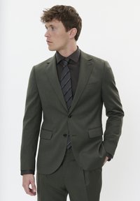 Tamsiai žalia kostiumas su liesu siluetu, vienas sagtys uždarymas, dvi priekinės kišenės, derinamas su tamsiai ruda elegantiška marškiniai ir plona dryžuota kaklaraiščiu.