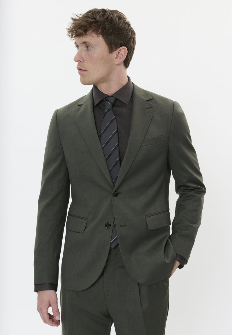 Tamsiai žalia kostiumas su liesu siluetu, vienas sagtys uždarymas, dvi priekinės kišenės, derinamas su tamsiai ruda elegantiška marškiniai ir plona dryžuota kaklaraiščiu.