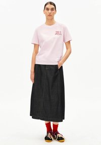 Hellrosa T-Shirt mit kurzen Ärmeln und rotem Text; dunkelblaue Maxirock aus Denim; rote Socken; schwarze Sneakers mit gelben Sohlen; Schnürsenkel und Akzente in Rosa.