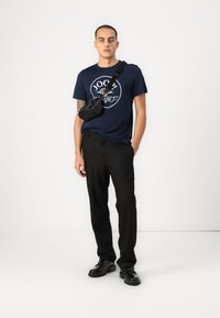 T-shirt bleu marine avec un imprimé graphique, pantalon noir et sac bandoulière noir. Des chaussures noires complètent cette tenue décontractée.