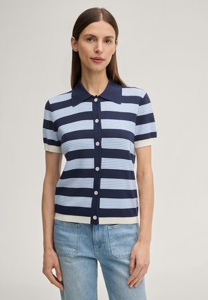 Kobieta ubrana w granatowo-niebieską, paskowaną koszulkę polo z krótkim rękawem zapiętą na guziki oraz jasne dżinsy, stojąca na jednolitym tle.