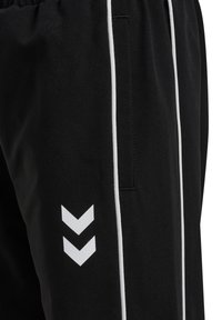 Sorte atletikbukser med hvid piping og to hvidt chevronlogoer pegende nedad nær lommen på låret.