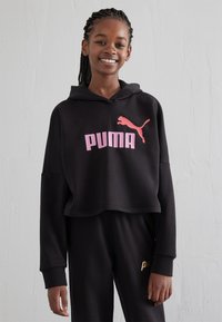 Sudadera corta negra de tejido suave, con un destacado logo de "PUMA" en rosa y una pequeña silueta de puma en rosa. Acompañada de pantalones negros.