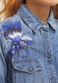 Denim shirt met knoopkraag en borstzak, met een gedetailleerd blauw en witte geborduurde vogel op de schouder.