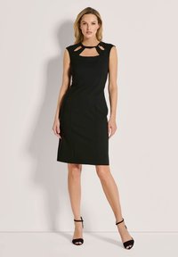 Schwarzes ärmelloses Kleid mit quadratischem Ausschnitt und Cut-out-Details. Figurbetontes Design, glatte Textur, knielang. Getragen mit schwarzen Absatzsandalen.