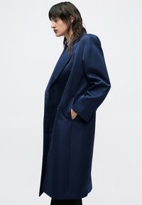 Tmavomodrý dlhý kabát so štruktúrovaným strihom, vyrobený z hladkej látky. Má široké klopy, bočné vrecká, priliehavý strih a jemné gombíky.