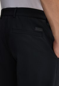 Pantalons noirs en tissu lisse, présentant une poche arrière avec une étiquette logo rectangulaire en relief et un design ajusté.