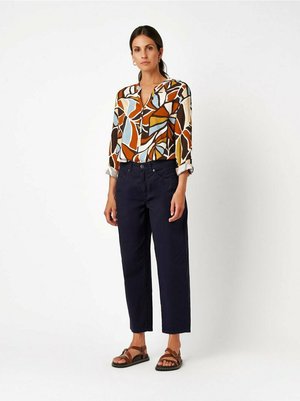 Woman with dark hair in a patterned blouse, navy high-waisted pants, and brown sandals standing against a white background.