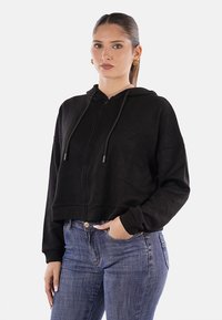 Felpa nera corta con maniche lunghe e cordini regolabili, dotata di una zip frontale e una texture morbida. Abbinata a jeans blu.
