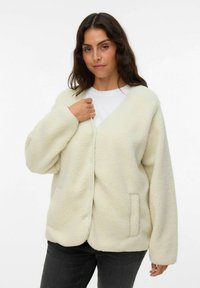 Cárdigan de crema con textura y esponjoso, con escote en V, hombros caídos y un bolsillo lateral. Fabricado con material sintético suave para proporcionar calor.