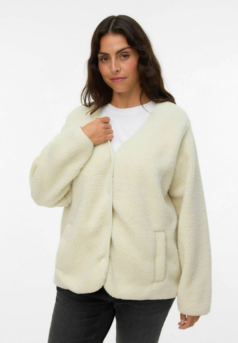Cárdigan de crema con textura y esponjoso, con escote en V, hombros caídos y un bolsillo lateral. Fabricado con material sintético suave para proporcionar calor.