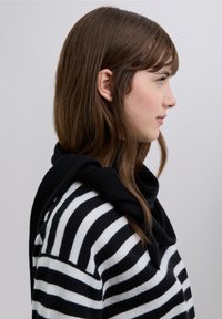 Svartvitrandig tröja med mjuk textur, lager på lager med en svart scarf som hänger runt halsen. Håret är långt och brunt.