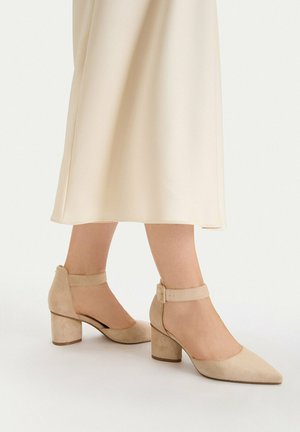 Beżowe zamszowe buty na słupku z zaostrzonymi noskami i paskami wokół kostki, noszone z kremową spódnicą midi na jasnym tle.