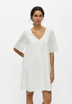 Jeune femme aux cheveux foncés en chignon bas, portant une robe blanche à texture florale, longueur genou, avec des manches aux coudes et un décolleté en V.