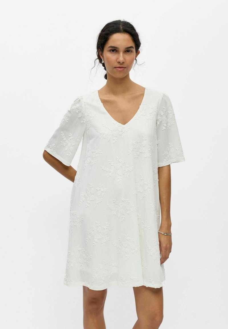 Jeune femme aux cheveux foncés en chignon bas, portant une robe blanche à texture florale, longueur genou, avec des manches aux coudes et un décolleté en V.