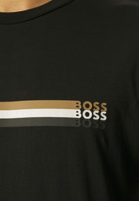Černé bavlněné tričko s horizontálními pruhy v zlaté, bílé a šedé, s nápisem "BOSS" vytištěným v různých barvách a tloušťkách.