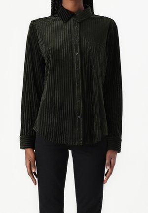 Person trägt ein dunkelgrünes Samthemd mit langen Ärmeln, vertikalen Streifen, durchgeknöpftem Vorderteil und Brusttasche, kombiniert mit schwarzen Hosen.