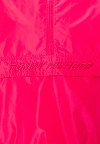 Wiatrówka w kolorze magenta o gładkiej, błyszczącej fakturze. Posiada zapięcie na zamek z przodu oraz haftowane logo "TOMMY HILFIGER" na dole.