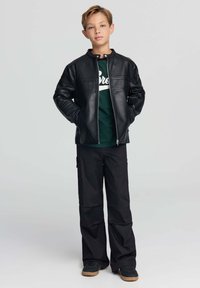 Ragazzo in piedi con le mani in tasca, indossa una giacca di pelle nera, una T-shirt verde con grafica, pantaloni neri larghi e sneaker nere con suola color gomma.