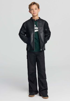 Ragazzo in piedi con le mani in tasca, indossa una giacca di pelle nera, una T-shirt verde con grafica, pantaloni neri larghi e sneaker nere con suola color gomma.