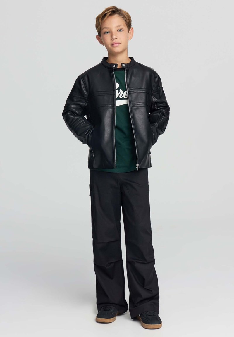 Ragazzo in piedi con le mani in tasca, indossa una giacca di pelle nera, una T-shirt verde con grafica, pantaloni neri larghi e sneaker nere con suola color gomma.