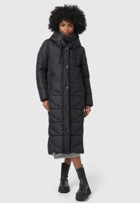 Long black puffer coat with a quilted pattern, adjustable hood, side pockets, and snap-button closure. Worn with black ankle boots.