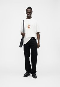 Man standing wearing white graphic t-shirt, black wide-leg pants, black shoes, and carrying black shoulder bag.