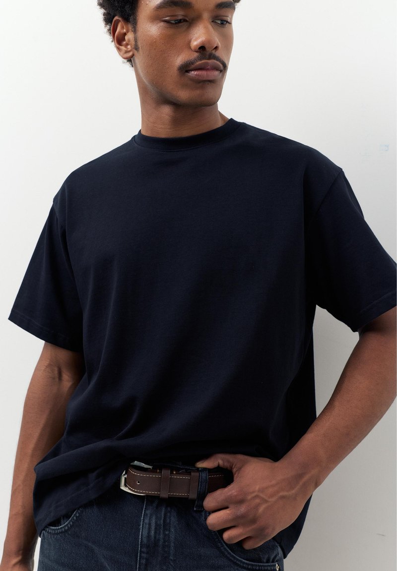 Junger Mann, der ein schlichtes schwarzes T-Shirt trägt, das in dunkle Jeans mit einem braunen Ledergürtel gesteckt ist, und seitlich blickt vor einem weißen Hintergrund.